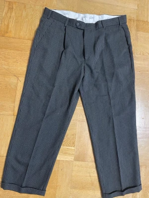 Randiga kostymbyxor från Simo Bellini - Snygga kostymbyxor från Simo Bellini i svart och grått med tunna vertikala ränder. Byxorna har klassisk passform, bälteshällor och är gjorda i en mix av 55% polyester och 45% ull. Perfekta för en stilren look.