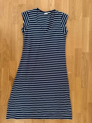 Boomerang - Marinblå randig kortklänning - Snygg marinblå kortklänning med vita horisontella ränder och v-ringad hals. Klänningen är ärmlös och har en avslappnad passform, perfekt för varma dagar. Tillverkad i mjukt bomullsmaterial för extra komfort.