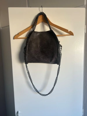 Mörkgrå axelväska med kedjedetaljer - Snygg mörkgrå axelväska i mockaimitation med coola kedjehandtag och lång axelrem i metall. Väskan har ett rymligt huvudfack och innerfack med dragkedja. Perfekt för dig som gillar en edgy och trendig look. Några flärpar har släppt ifrån kedjan men det påverkar inte funktionen av väskan alls