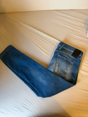 Blå raka jeans från Replay - Säljer ett par klassiska blå jeans från Replay med raka ben och femficksmodell. Jeansen har en snygg tvätt och ett svart läderpatch med logga bak i midjan. Perfekta till vardagslooken och tillverkade i slitstark denim.
