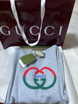 Grå hoodie från Gucci med logga - Snygg grå hoodie från Gucci med stor färgglad logga i rött, grönt och blått på bröstet. Tillverkad i mjuk bomull och har klassisk passform med ribbade muddar. Perfekt för dig som vill ha en exklusiv och trendig look. Allt följer med den fina hoodie.