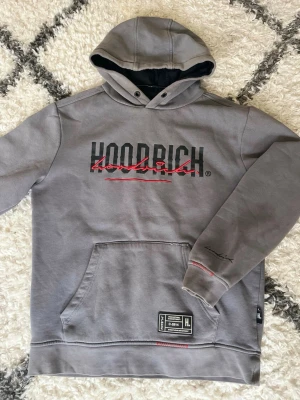 Grå hoodie från Hoodrich - Snygg grå hoodie från Hoodrich med svart och rött tryck på bröstet och en stor magficka. Detaljerad etikett nedtill och broderad logga på ärmen samt gömd liten ficka i ärmen. Perfekt för en avslappnad streetwear-look.