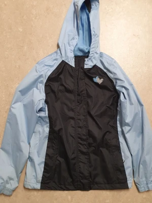 Vindjacka - Snygg vindjacka i ljusblått och svart med huva och dragkedja framtill. Jackan har kardborrestängning över dragkedjan och ett litet hjärtmärke på bröstet. Tillverkad i tunt, vindtätt material som passar perfekt för blåsiga dagar.