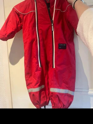 Röd vindoverall från Polarn O. Pyret - Snygg röd vindoverall från Polarn O. Pyret med vita dragkedjor, reflexdetaljer och resår vid benslut. Overallen har en klassisk design och är tillverkad i slitstarkt material som står emot vind. Perfekt för blåsiga dagar och utomhuslek.