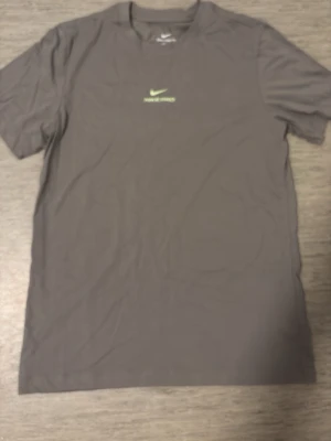 Nike Pro grått träningsset - Säljer ett grått träningsset från Nike Pro med t-shirt och shorts. Båda plaggen har Nike-logga och text i ljusgrönt framtill. T-shirten har klassisk passform och shortsen har bred resår med Nike Pro-tryck. Perfekt för gym eller löpning.