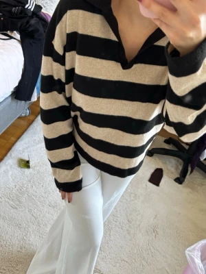 Randig tröja med krage svart/beige - Säljer en oversized tröja med breda svarta och beige ränder samt krage. Tröjan har långa ärmar och en avslappnad passform, perfekt för en chill look. Materialet känns mjukt och bekvämt mot huden.