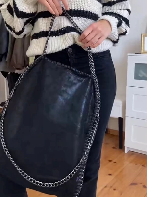Stella Mcarntby västa! - Stilren svart axelväska i glansigt material med kraftiga silverfärgade kedjor som handtag och kantdetalj. Väskan har en mjuk, slouchy form och rymmer mycket. Perfekt för dig som gillar en edgy och trendig look.Inte äkta ❤️