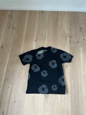 Svart t-shirt med blommigt tryck - Snygg svart t-shirt från denim tears med stora gråa blommor tryckta över hela tröjan. Klassisk passform med rund hals och korta ärmar. Perfekt för dig som vill ha en unik och trendig look.