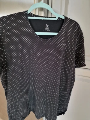 Svart prickig t-shirt. - Svart t-shirt från Eco Ica maxi med små vita prickar över hela plagget. Klassisk rund halsringning och korta ärmar. Tillverkad i mjuk bomull som känns skön mot huden. Perfekt för en avslappnad och stilren look. I