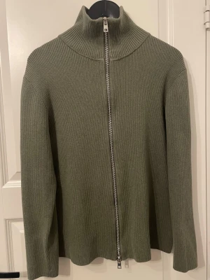 Olivgrön stickad tröja med dragkedja - Snygg olivgrön stickad tröja från Ljung by Marcus Larsson. Tröjan har hög krage och hel dragkedja framtill. Tillverkad i ribbstickat material som ger en stilren och modern look. Perfekt för lager på lager och enkel att matcha.