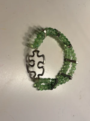 Grönt pärl armban med små diamanter och pusselbits bricka  - Snyggt armband med tre rader gröna pärlor och en unik metallbricka i form av en pusselbit. Armbandet har små metallspännen som håller ihop pärlraderna och ger en cool touch. Perfekt accessoar för dig som gillar färg och detaljer.