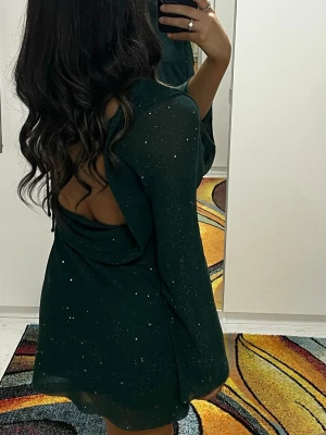 Mörkgrön glittrig klänning med öppen rygg - Snygg mörkgrön kortklänning med glittriga detaljer och öppen rygg. Klänningen har långärmade ärmar och en figurnära passform. Perfekt för dig som vill sticka ut med en elegant look. Materialet känns mjukt och stretchigt.