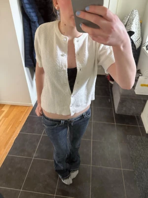 Vit kortärmad kofta med knappar - Söt vit kofta med korta ärmar och rund hals. Koftan har knäppning framtill med små vita knappar och ribbade kanter vid ärmslut och nederkant. Perfekt att slänga över ett linne eller topp för en enkel och fräsch look.