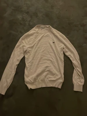 Vit/tröja från Lacoste - Klassisk vit långärmad tröja från Lacoste med rund halsringning och den ikoniska krokodilloggan på bröstet. Tröjan har ribbade muddar vid ärmslut och nederkant, och är tillverkad i ett mjukt och tunt material som passar perfekt till lager på lager.
