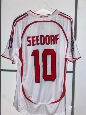 AC Milan Seedorf 10 bortatröja 2007 - Säljer en vit AC Milan bortatröja från Champions League-finalen 2007 med Seedorf och nummer 10 på ryggen. Tröjan har röda och svarta detaljer, klubbmärke, Adidas-logga och Champions League-patch på ärmen. Tillverkad i lätt och ventilerande material.