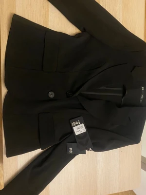 Svart kavaj från Prett - Stilren svart kavaj från Prett med klassisk passform och en knapp framtill. Kavajen har två framfickor och är tillverkad i ett slätt tyg som ger en snygg siluett. Perfekt för dig som vill ha en enkel men trendig look.