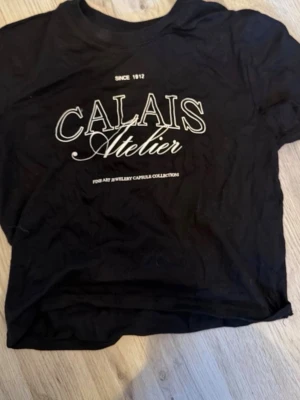 Svart crop top CALAIS Atelier - Svart crop top t-shirt från CALAIS Atelier med vit text framtill. T-shirten har rund halsringning och korta ärmar. Materialet ser ut att vara mjukt och stretchigt, perfekt för en avslappnad och trendig look.