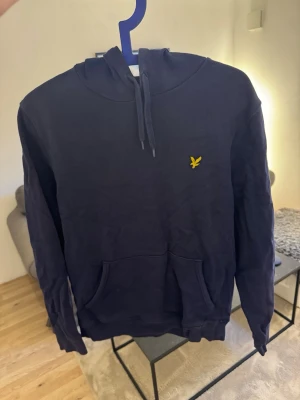 Mörkblå hoodie från Lyle & Scott - Snygg mörkblå hoodie från Lyle & Scott med klassisk gul logga på bröstet. Hoodien har huva med dragsko och en stor magficka framtill. Tillverkad i mjukt bomullsmaterial och har ribbade muddar vid ärmslut och nederkant. Perfekt för en avslappnad stil.