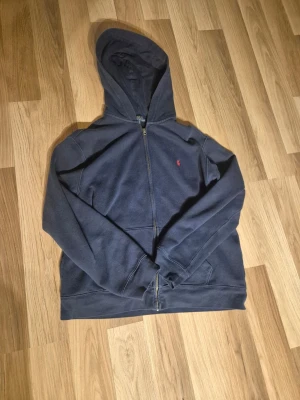 Mörkblå zip hoodie från Polo Ralph Lauren - Mörkblå hoodie med dragkedja från Polo Ralph Lauren. Klassisk design med huva, fickor framtill och den ikoniska lilla röda logon broderad på bröstet. Perfekt för en avslappnad och stilren look. Mjuk bomullsblandning som känns skön mot huden.