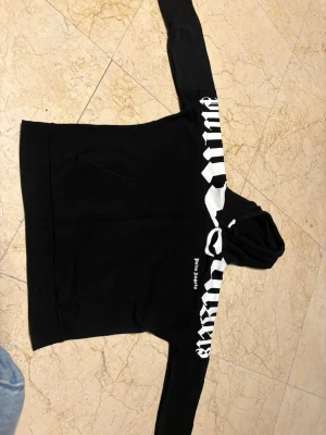 Svart hoodie från Palm Angels - Snygg svart hoodie från Palm Angels med vit grafisk text över bröstet och ena ärmen. Klassisk känguruficka framtill och huva. Tillverkad i mjukt material som känns skönt mot huden. Perfekt för en streetwear-look.