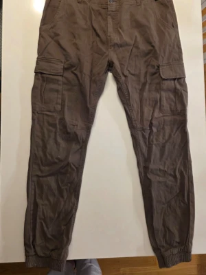 Olivgröna cargobyxor med muddar - Säljer ett par bruna cargobyxor med stora fickor på sidorna och muddar vid bensluten. Byxorna har en avslappnad passform och är gjorda i ett mjukt bomullsmaterial. Perfekta för dig som gillar en chill och praktisk stil.      Midja:  44cm    Benlängd: 75 cm          Hela: 105cm