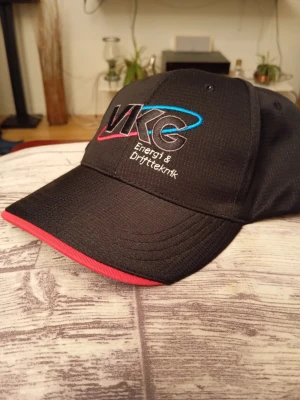 Ny Svart keps med broderad logga - Snygg svart keps med böjd skärm och broderad VKG Energi & Driftteknik-logga i vitt, blått och rosa framtill. Skärmen har en cool röd kant på undersidan. Perfekt för dig som vill ha en unik och stilren accessoar.