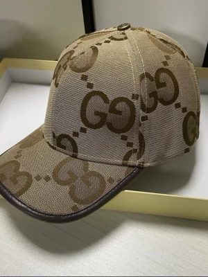 Beige keps med GG-mönster från Gucci - Säljer en beige keps från Gucci med det klassiska GG-mönstret i brunt över hela kepsen. Kepsen har böjd skärm med mörkbrunt kantband och är tillverkad i canvasmaterial. Perfekt för dig som vill ha en stilren och lyxig accessoar.