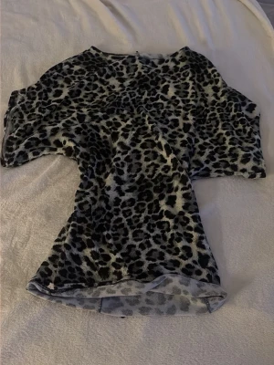 Leopardmönstrad topp med kort ärm - Säljer en snygg topp med leopardmönster i svart och grått. Tajt vid midjan och lös uppe. Toppen har korta ärmar och en rund halsringning. Materialet känns mjukt och stretchigt, perfekt för dig som vill sticka ut lite extra.