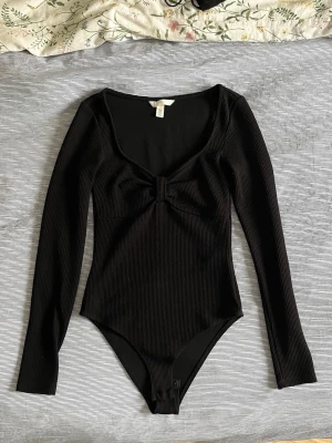 Svart ribbad body med rosett - En svart långärmad ribbad body med låg urringning och en rosettdetalj på bröstkorgen. Knäppning nere på bodyn för lätt av och påtagning. 