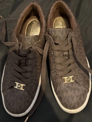 Bruna Michael Kors sneakers - Snygga bruna sneakers från Michael Kors med diskret logomönster över hela skon och guldfärgade MK-detaljer på snörningen. Skorna har vita sulor och är tillverkade i ett tåligt syntetmaterial med textilkänsla. Perfekta för dig som gillar stilrena och trendiga skor.