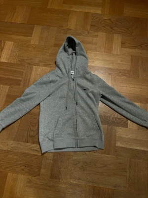Grå Lacoste hoodie (HELT NY) - Säljer en grå Lacoste zip hoodie då den var för liten för mig. Helt ny och aldrig använd förutom när jag provat den och fotat den. Storlek S. Nypris ca 1300kr. Säljer för 400kr.