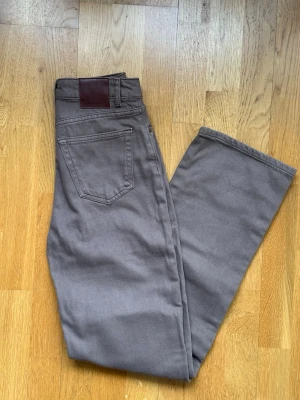 Bruna jeans från Never Denim - Säljer ett par bruna jeans från Never Denim med klassisk femficksdesign och rak passform. Jeansen har en snygg läderpatch Bruna jeans från Never Denim i midjan och är tillverkade i ett slitstarkt bomullsmaterial. Perfekta för dig som gillar en enkel och stilren look.