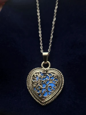 Halsband med hjärthänge i silverfärg - Säljer ett helt unikt, handgjort halsband i silverfärgat stål. Hänget är ett vackert ornamenterat hjärta med snirkliga detaljer och en dekorativ kant, ett riktigt statement-smycke! Det magiska: Efter att ha varit i vanligt dagsljus en liten stund, börjar hjärtat lysa i en förtrollande blå färg i mörkare omgivning. Ju mer ljus det får, desto starkare och längre lyser det (upp till hela 12 timmar!). Detaljer: Kedjan är 45 cm, tunn och justerbar i rostfritt stål. Hänget är ca 3 cm.