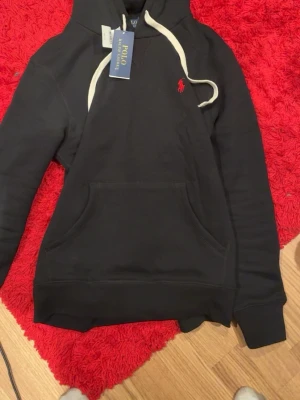 Svart hoodie från Polo Ralph Lauren - Snygg svart hoodie från Polo Ralph Lauren med vit dragsko i huvan och röd broderad logga på bröstet. Klassisk känguruficka framtill och mjukt material som känns skönt mot huden. Perfekt för en avslappnad och stilren look.