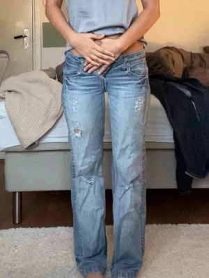 Ljusblå bootcut jeans med slitningar - Säljer ett par ljusblå straight jeans med slitningar och hål på benen. Jeansen har coola broderade detaljer på bakfickorna och klassisk femficksmodell. Tillverkade i denim med en avslappnad passform och låg midja. inerbenslängd:74cm  midja tvärs över: 36cm 