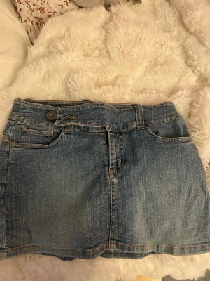 Blå lågmidjad jeanskjol med fickor - Säljer en klassisk blå jeanskjol i kort modell med fem fickor och knapp- samt dragkedjestängning framtill. Kjolen har bälteshällor och är gjord i ett slitstarkt denimtyg som passar perfekt till många olika stilar.