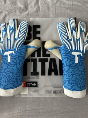 Blå målvaktshandskar från T1TAN - Säljer ett par blå målvaktshandskar från T1TAN med vitt mönster och vita detaljer. Handskarna har greppvänlig latex på handflatan och elastisk mudd med texten 'BE THE TITAN'. Perfekta för fotbollsträning eller match. Handskarna har bara blivit använd 3 gånger och de är själv designade. Priset går att diskutera.