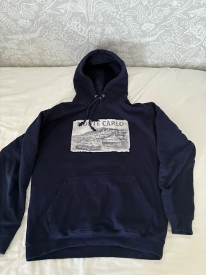 Monte Carlo Hoddie - Snygg mörkblå hoodie med stor ficka framtill och dragsko i huvan. På bröstet finns ett stort tryck med texten 'Monte Carlo' och motiv av en bil, båt och stadsmiljö. Perfekt för dig som gillar streetwear och vill ha en unik look.