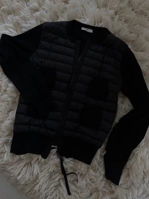 Svart cardiganjacka från Moncler - Snygg svart cardiganjacka från Moncler med quiltad front och stickade ärmar. Jackan har dragkedja framtill, ribbade muddar och fyra fickor. Perfekt för lager på lager och en stilren look.