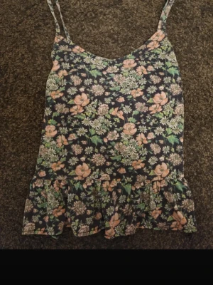 Blommigt linne - Söt linne med smala axelband och volangkant nedtill. Toppen har ett blommigt mönster i grönt, rosa och vitt på mörk botten. Perfekt för varma dagar och enkel att matcha med jeans eller kjol! Jöttesnygg