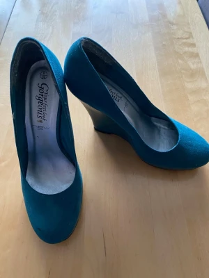 Turkosa kilklackade pumps Gorgeous - Snygga turkosa pumps från Gorgeous med kilklack och rundad tå. Skorna har en klassisk siluett och är tillverkade i textilmaterial som ger en mjuk känsla. Perfekta för dig som vill ha en färgklick till outfiten.