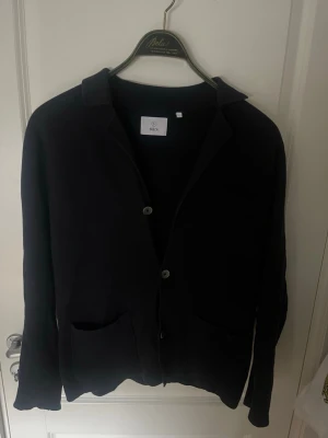 Mörkblå kofta från Bläck med knappar - Stilren mörkblå kofta från Bläck med knäppning framtill och klassisk krage. Perfekt att slänga över en t-shirt eller skjorta för en clean look. Långärmad och mjukt material som känns skönt mot huden.