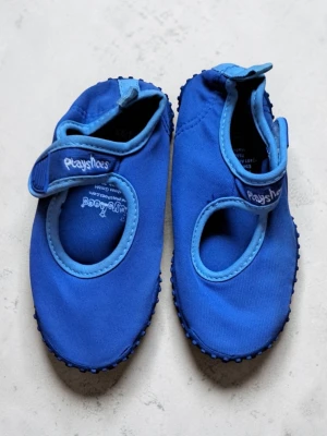 Blå badskor från Playshoes - Säljer ett par blå badskor från Playshoes med kardborreband och rundad tå. Skorna har en mjuk ovandel i textil och en flexibel sula med små gummiprickar för extra grepp. Perfekta för badsäsongen, inomhus och utomhus. 