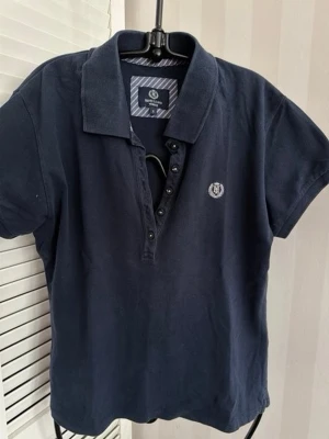 Mörkblå pikétröja från Henry Lloyd  - Klassisk mörkblå pikétröja från Henry Lloyd med korta ärmar och krage. Tröjan har knappar framtill och en liten broderad logga på bröstet. Tillverkad i mjuk bomull som känns skön mot huden.Storlek M-L