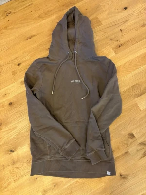 Brun hoodie från Les Deux - Säljer en brun hoodie från Les Deux med broderad logga på bröstet. Hoodien har dragsko i huvan och en klassisk känguruficka framtill. Tillverkad i mjukt bomullsmaterial och har ribbade muddar vid ärmslut och nederkant. Perfekt för en avslappnad och stilren look.