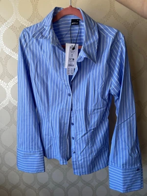 Blåvit randig skjorta från Ginatricot - Snygg blå skjorta med vita ränder från Ginatricot. Skjortan har långa ärmar, klassisk krage, knappar framtill och knytband i ryggen för en mer figurnära passform. Tillverkad i bomull, perfekt för en fräsch och stilren look. Har aldrig använt den så den är ny! 