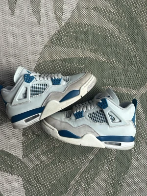 Air Jordan 4 Retro military blue  - Säljer ett par Air Jordan 4 Retro sneakers i vitt och blått med meshpaneler och detaljer i plast. Skorna har en chunky sula och ikoniska vingar på sidorna. Perfekta för dig som gillar klassisk streetwear och vill sticka ut med en tidlös modell.