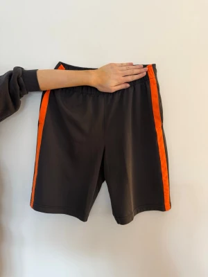 Mörkgrå shorts XS - Säljer ett par mörkgrå shorts med orangea ränder längs sidorna. De har elastisk midja och är tillverkade i ett lätt och sportigt material. 