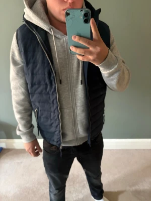 Väst med huva - Säljer en mörkblå quiltad dunväst med huva och dragkedja framtill. Västen har en clean look med fickor på sidorna och passar perfekt över en hoodie för en avslappnad stil. Perfekt för kyliga dagar när du vill hålla värmen utan att bära jacka.