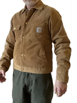 Brun manchesterjacka från Carhartt - Snygg brun manchesterjacka från Carhartt med klassisk krage, knappstängning och bröstficka med logopatch. Jackan har raka linjer och en avslappnad passform, perfekt för dig som gillar streetwear och tidlös stil.